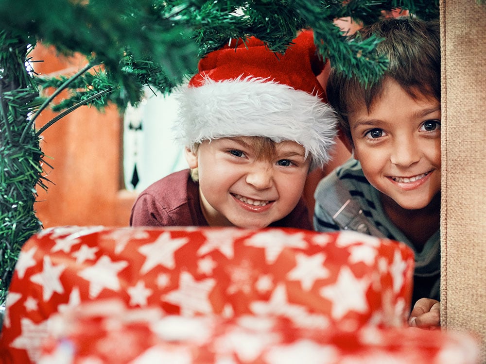 Siblings under a Christmas tree grinning at presents.